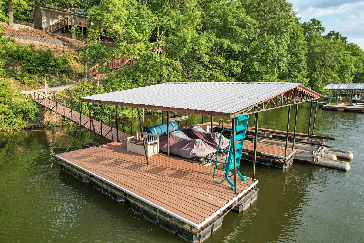The image features a peaceful lakeside scene with a charming boathouse, a spacious dock equipped for boating and fishing, and calm water reflecting the natural wooded surroundings. The dock includes boat covers and a ladder, inviting guests to relax by the water, launch a boat for a day on the lake, or enjoy the view as the sun sets over the Ozarks.
