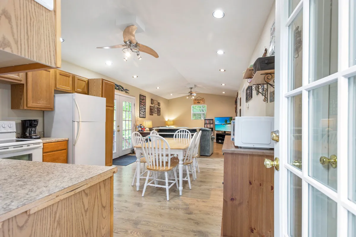 A warm and inviting open-concept space combining the kitchen, dining table, sofa, and a visible television. Decorative wall art adds personality, while large windows and a glass door allow natural light to flood the area, hinting at beautiful lake views just beyond.