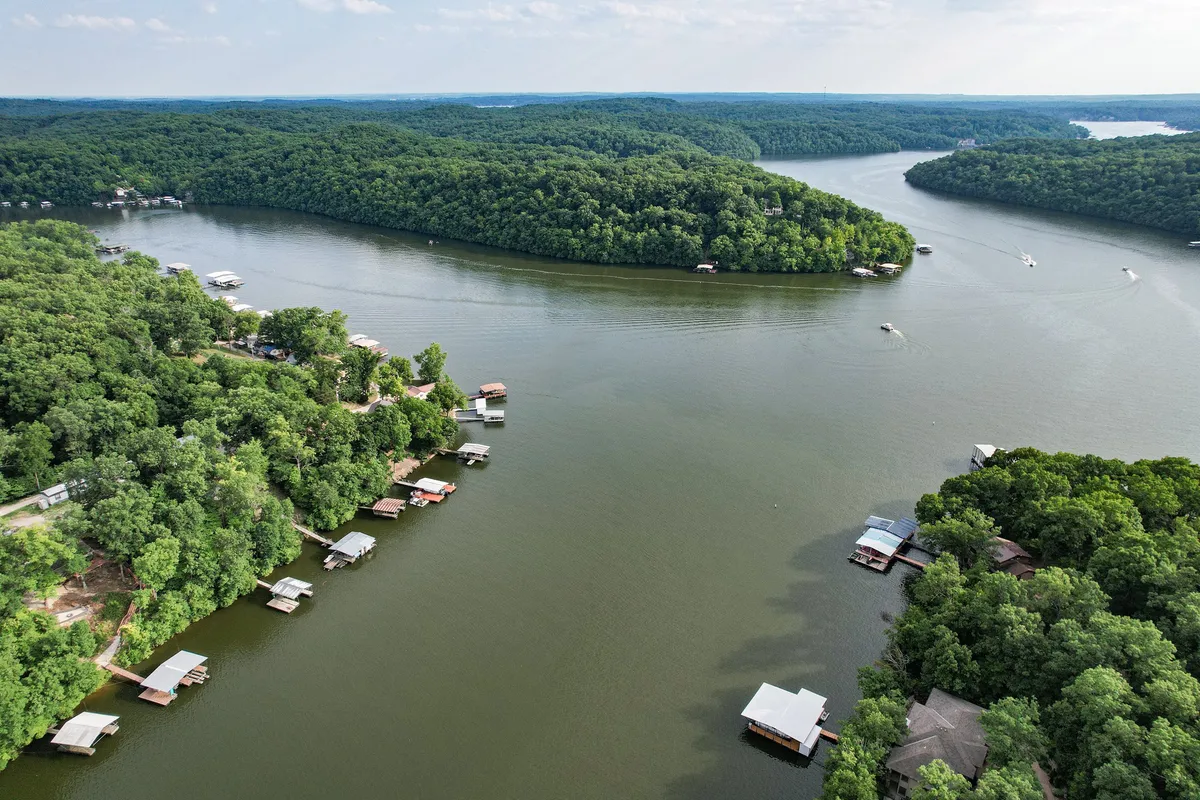 A panoramic view of lush green Ozark hills, a serene waterway bordered with boathouses, and distant islands. The scene captures the natural landscape that embodies local exploration and outdoor adventure.