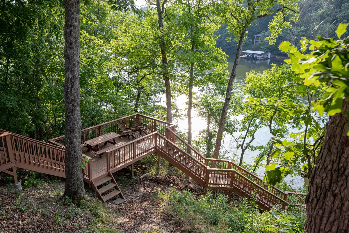 A wide wooden deck features a picnic table and stairs leading down to the lake, framed by dense trees. The lake is calm and inviting, and the natural surroundings create a peaceful, immersive experience with opportunities for wildlife watching and lakeside activities.