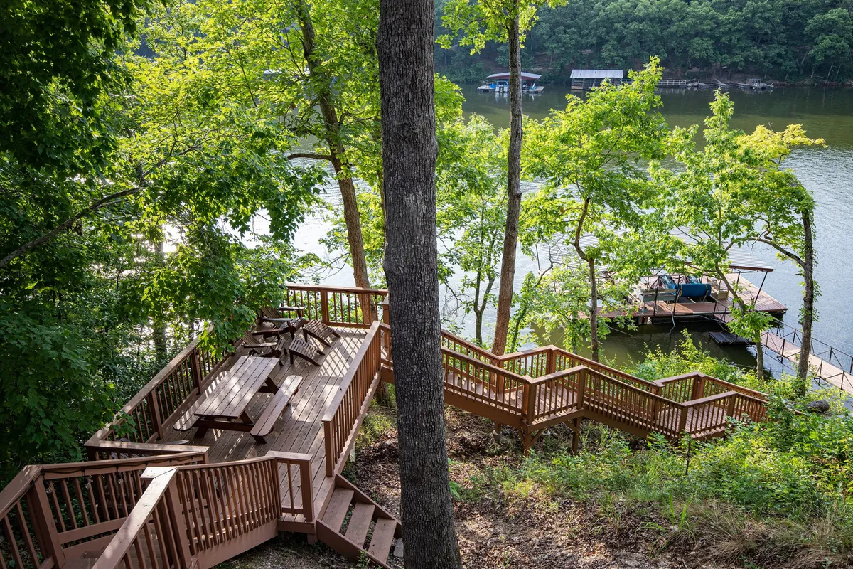 A broad wooden deck overlooks the tranquil lake, framed by a staircase leading down through the trees. The scene is set for serene mornings and peaceful evenings, with nature enveloping the space and the shimmering water visible beyond the deck railing.