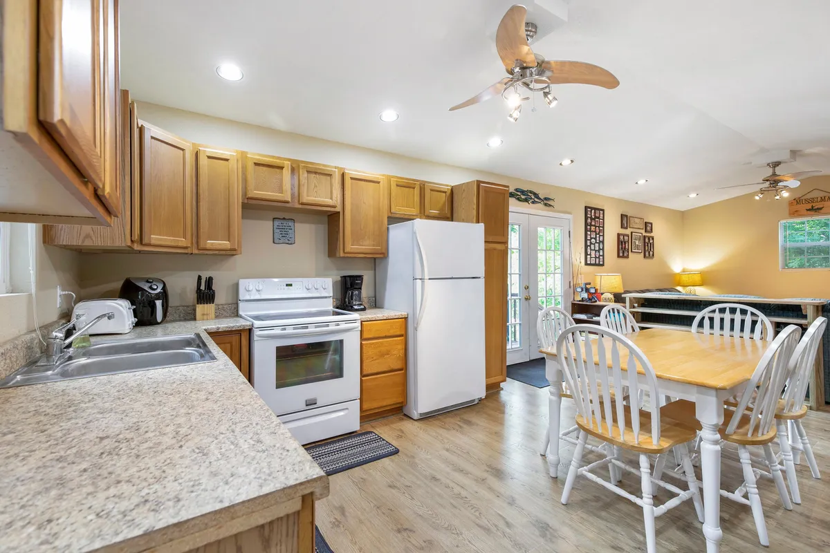 A spacious kitchen and dining area featuring a large window that frames the outdoors and fills the room with golden light. The kitchen island and dining table invite shared meals and socializing, while the modern yet rustic décor echoes the lakehouse theme.