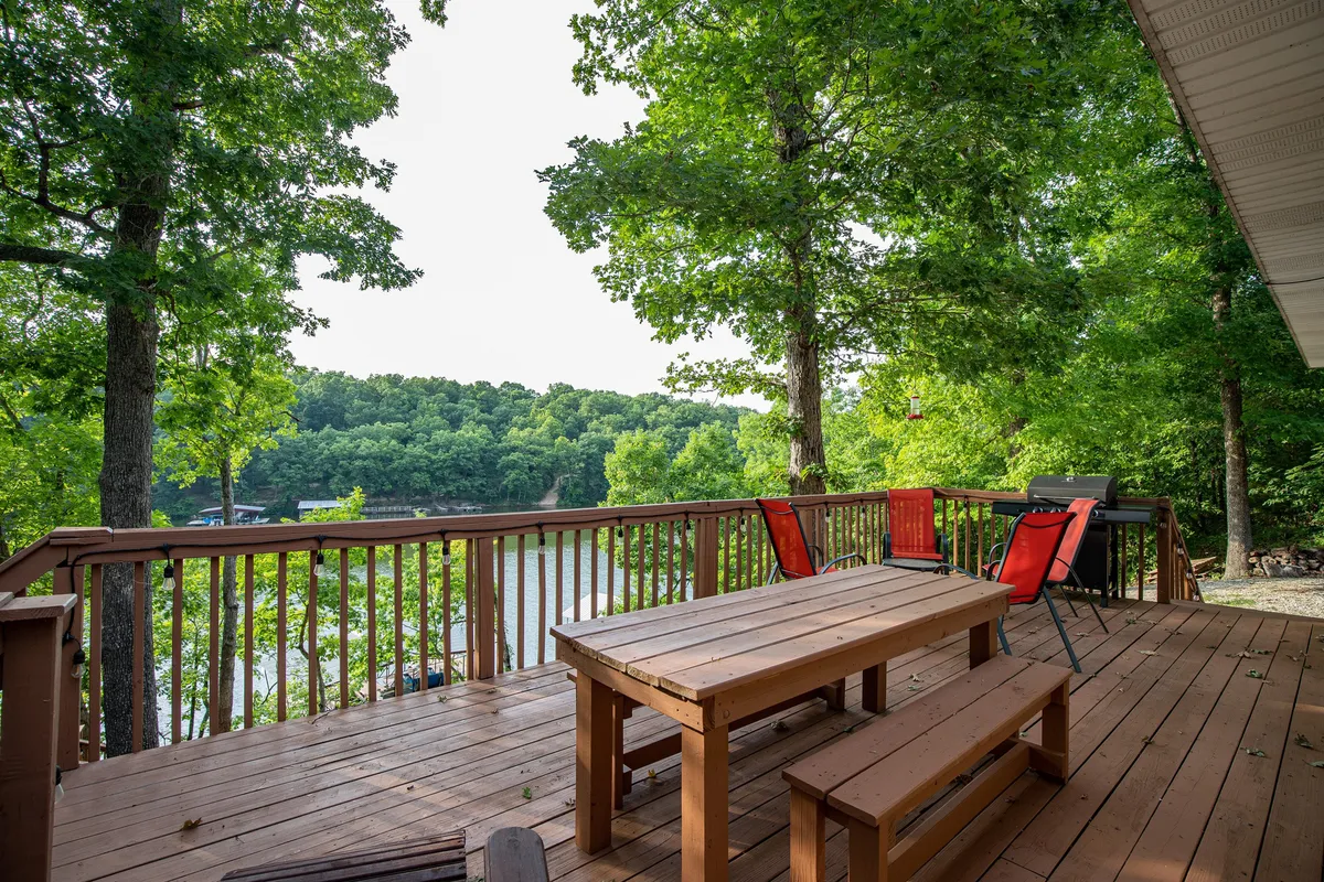 A tranquil wooden deck with red chairs overlooks the serene lake, surrounded by tall trees. The view is peaceful, with no signs of boat traffic, and the deck invites moments of relaxation, outdoor dining, or simply soaking in nature. The distant shoreline, gentle water, and lush greenery emphasize the secluded atmosphere.