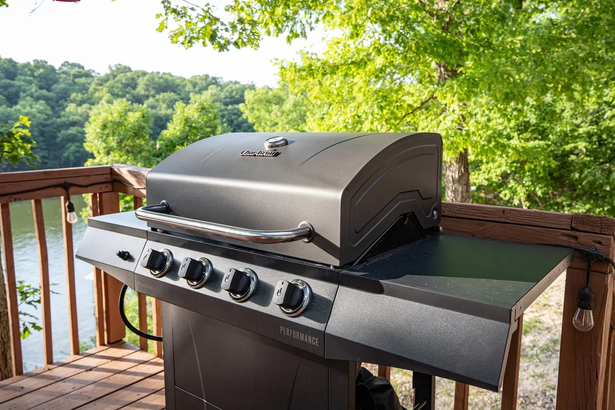An outdoor scene highlighting a grill stationed on a deck overlooking lush greenery and water. The background hints at the serene lakeside environment while foreground features invite guests to socialize and dine al fresco.