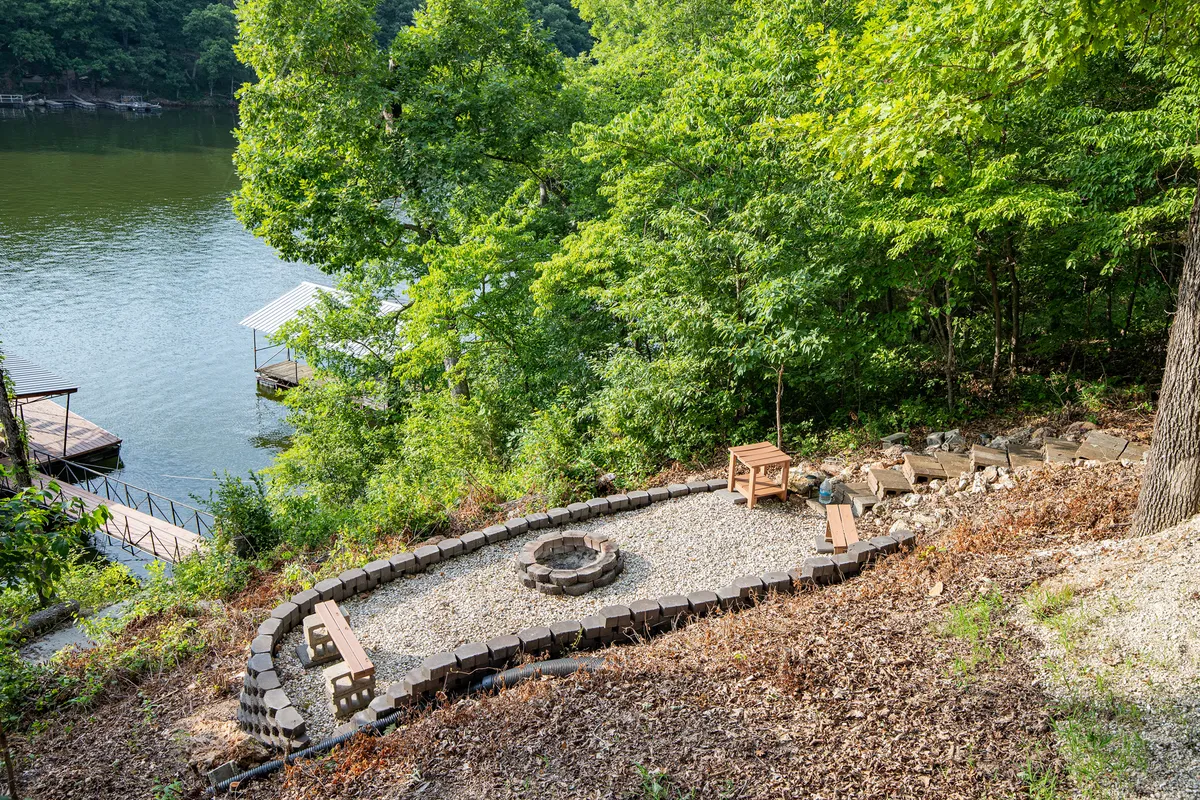 A cozy fire pit with surrounding seating sits nestled among natural foliage, with the lake shimmering just beyond. The arrangement invites evening gatherings under the stars, embodying the campfire experience described.