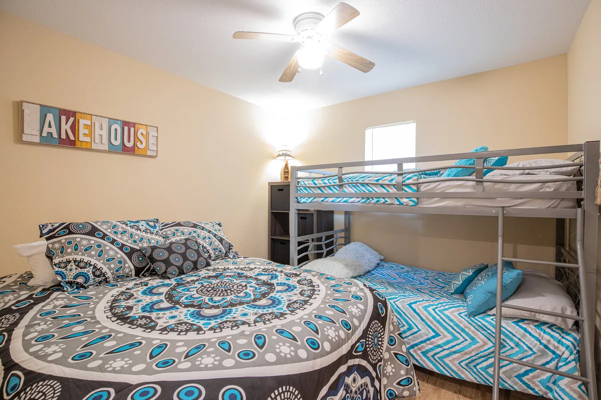 This interior image displays a bedroom featuring both a queen bed and a twin-sized bunk bed, with accent pillows and playful lakehouse decor. The arrangement demonstrates the room’s versatility and the way sleeping spaces are maximized for comfort and group stays.